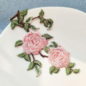 Puede incluir: Apliques florales bordados con rosas rosas y hojas verdes sobre tallos marrones. El bordado detallado crea un efecto texturizado. Ideales para embellecer ropa, accesorios o proyectos de manualidades.
