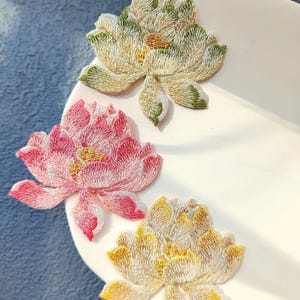 May include: Three embroidered lotus flower patches in cream, pink, and yellow. The flowers are detailed with green and gold accents, and are arranged on a white, curved surface. The patches are likely for decorative use on clothing or accessories.