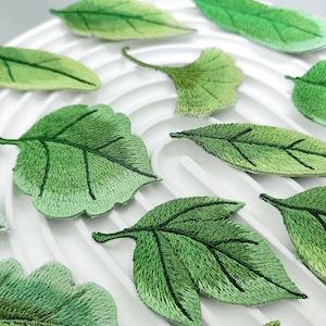 Può includere: Una collezione di toppe ricamate a forma di foglia in varie tonalità di verde, con cuciture dettagliate che imitano le venature naturali. Le toppe sono disposte su una superficie bianca con scanalature circolari concentriche.