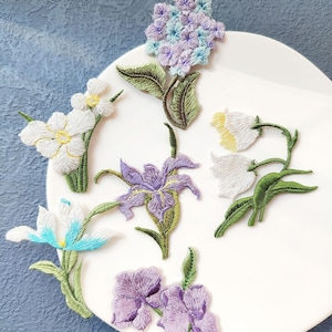 May include: Embroidered floral patches in various colors, including white, purple, and blue, are arranged on a white plate. The patches depict different flower types, such as irises and hydrangeas, with green leaves and stems. The embroidery details are visible.