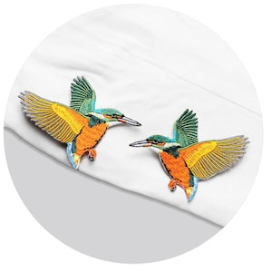 Op de afbeelding: Twee geborduurde strijk-op patches van ijsvogels. De vogels kijken in tegengestelde richtingen en hebben groene, blauwe, gele en oranje details.