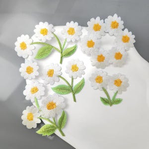 Puede incluir: Una colección de parches bordados de margaritas con pétalos blancos, centros de color amarillo brillante y hojas de color verde claro. El conjunto incluye flores individuales y pequeños grupos con tallos, realizados con hilo detallado.