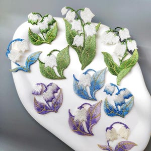 以下が含まれることがあります： 様々な色のスズランの刺繍パッチ。白いベル型の花と緑の葉、青と紫のバリエーションが特徴です。パッチは白い表面に配置されています。