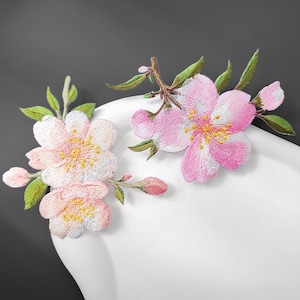 May include: Two embroidered cherry blossom patches featuring pink and white petals with yellow centres, green leaves, and small buds on brown stems. The embroidery displays detailed stitching and vibrant colours, showcasing floral branches.