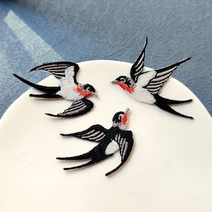 May include: Three embroidered swallow patches. Each patch features a black body, white chest, and red throat. The swallows have outstretched wings, and are arranged on a white surface.