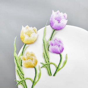 May include: Two embroidered tulip patches featuring detailed stitching. One patch displays two yellow tulips with green stems and leaves, while the other patch shows two purple tulips with green stems and leaves, all rendered in vibrant thread colors.