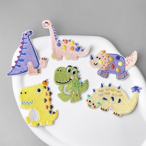 Puede incluir: Un conjunto de seis parches bordados de dinosaurios que incluye un dinosaurio de cuello largo morado, uno rosa, un dinosaurio verde de pie, uno amarillo, un triceratops morado y un anquilosaurio amarillo con detalles coloridos.