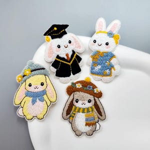 May include: Five embroidered bunny patches in different outfits. One bunny is in a graduation cap and gown, others wear hats and scarves. The bunnies are white, yellow, and brown, with colourful accents. These patches are ideal for adding a whimsical touch to clothing or accessories.