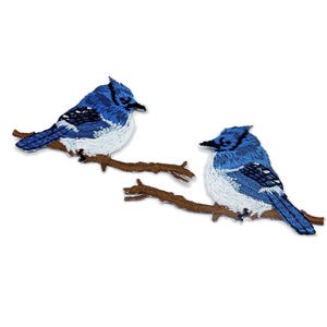 Op de afbeelding: Twee geborduurde patches van blauwe gaaien op bruine takken. De vogels hebben blauwe en witte veren met zwarte snavels. De patches staan op een witte achtergrond.