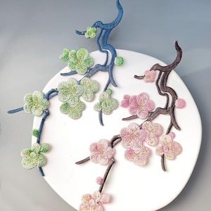 May include: Embroidered floral branch designs in green, pink, and blue. The floral branches are arranged on a white circular background. The flowers have detailed stitching and a textured appearance, creating a three-dimensional effect.