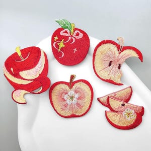 May include: A collection of five embroidered apple patches in various forms, including a whole apple with a bow, a peeled apple, a sliced apple, and two apple wedges. The designs feature vibrant red, yellow, and cream thread colors with detailed stitching.