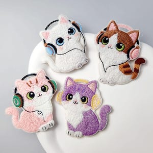 May include: Four embroidered cat patches in white, pink, purple, and brown, each wearing headphones. The cats have large eyes and are sitting or reclining. The patches are on a white surface.