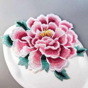 Puede incluir: Parche floral bordado con una gran peonía con pétalos rosas y blancos, centro amarillo y hojas verdes. El parche es un diseño detallado y colorido, adecuado para embellecer ropa o accesorios.