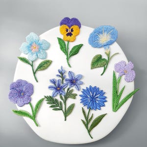 May include: A collection of seven embroidered flower patches featuring various blue, purple, and yellow floral designs with green stems and leaves. Each patch displays intricate thread work and distinct petal shapes, arranged on a white circular base.