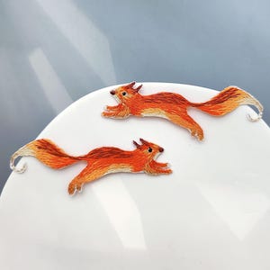 Peut inclure: Deux appliqués brodés représentant des écureuils roux et bruns en mouvement. Les écureuils sont détaillés avec du fil, mettant en valeur leur fourrure et leurs caractéristiques. Les appliqués sont posés sur une surface blanche.