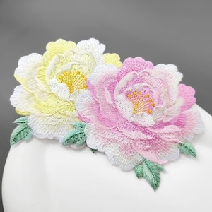 以下が含まれることがあります： 2つの大きな重なり合う牡丹の花が刺繍されたワッペン。1つの花は白のハイライトが入った淡い黄色で、もう1つは白の縁取りがあるソフトピンクです。どちらの花も中心が黄色で、小さな緑色の刺繍の葉がアクセントになっています。