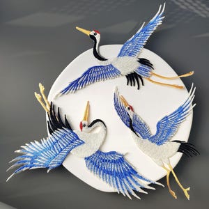 Può includere: Opera d'arte ricamata con tre gru in volo. Le gru hanno piumaggio bianco, nero e blu con accenti rossi sulla testa e becchi e zampe gialle. L'opera è montata su una base circolare bianca.