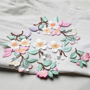 Può includere: Applique floreali ricamate con fiori bianchi, rosa e gialli, foglie verdi e steli marroni. Il design floreale è posto su uno sfondo di tessuto bianco, creando un'estetica delicata e dettagliata.