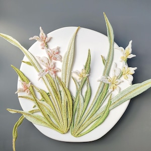 May include: A white circular wall art piece featuring embroidered flowers and leaves. The artwork showcases delicate pink, white, and yellow blossoms with green and gold-toned leaves. The design is set against a gray background, highlighting the intricate details.