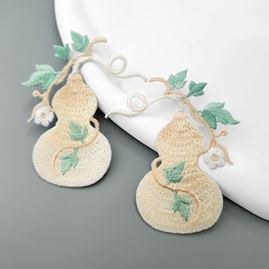 以下が含まれることがあります： ベージュ色のひょうたんの形をした刺繍ワッペン2枚。薄緑色の葉、カールした蔓、小さな白い花が刺繍されています。刺繍には細かなステッチが施されており、それぞれのひょうたんの表面に質感のある外観を作り出しています。
