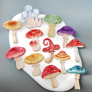 Könnte beinhalten: Eine Sammlung von gestickten Pilzaufnähern in verschiedenen Farben, darunter Rot, Gelb, Grün, Blau und Lila. Die Aufnäher haben detaillierte Stickereien und sind auf einer weißen, geschwungenen Oberfläche angeordnet. Die Pilze haben unterschiedliche Formen und Größen.