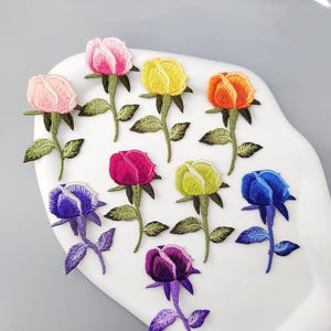 May include: A collection of nine embroidered rose patches in various colors, including pink, yellow, orange, red, green, blue, and purple. Each patch features a detailed rose bloom, green leaves, and a stem. The patches are arranged on a white, curved surface.