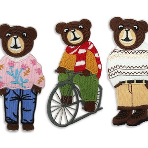 May include: Three embroidered iron-on patches featuring brown bears wearing different outfits. One bear is wearing a pink sweater with floral designs and blue jeans. Another bear is wearing a yellow sweater with a red and white striped scarf and green pants, riding a bicycle. The third bear is wearing a white sweater with brown and white stripes and beige pants.