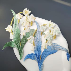 May include: Embroidered floral appliques featuring white bell-shaped flowers with yellow accents, green leaves, and blue leaves. The appliques are on a white, curved surface, creating a decorative floral design.