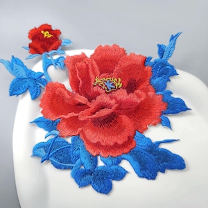 Puede incluir: Un parche bordado que presenta una gran peonía de color rojo vibrante con un centro amarillo, rodeada de hojas y tallos de color azul brillante. Un capullo de peonía rojo más pequeño y a juego se sitúa arriba a la izquierda de la flor.