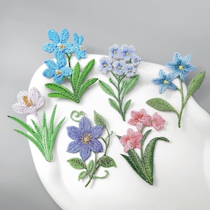 Könnte beinhalten: Eine Sammlung von sechs gestickten Blumenaufnähern mit verschiedenen botanischen Motiven. Das Set enthält hellblaue, blaue, violette und rosafarbene Blüten mit detaillierten grünen Blättern und Stielen, alles in farbigem Garn auf weißem Grund.