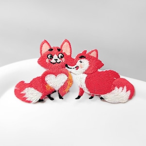 May include: Two embroidered iron-on patches featuring stylised foxes in shades of red, white, and black. The left fox has a white heart shape on its chest, while the right fox is positioned to the side, both with detailed stitching and pointed ears.