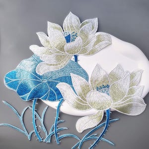 Può includere: Fiori di loto ricamati in bianco e verde con dettagli blu. Il design include una foglia blu e steli con spine. I fiori sono ambientati su uno sfondo bianco curvo, il tutto su uno sfondo grigio. Il ricamo è dettagliato e artistico.