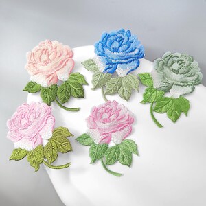 以下が含まれることがあります： 詳細な花びらと緑の葉が特徴の5つの刺繍入りローズワッペン。花はライトピンク、ブライトブルー、ペールグリーン、ミディアムピンク、ダークピンクの色合いで、それぞれ独特のステッチと重なり合った花のような質感が施されています。