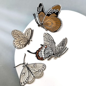 May include: Four embroidered butterfly patches in various colours and patterns. One butterfly has brown wings with black and white accents, while others feature shades of grey and tan. The patches are arranged on a white surface.