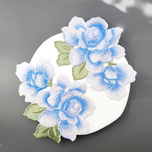 May include: Three embroidered floral patches featuring layered petals in shades of white and bright blue. Each flower is accented with light green leaves with visible stitching detail. The patches are arranged on a circular white surface.