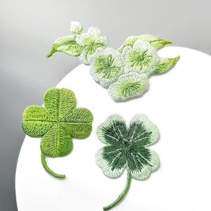 May include: Three embroidered patches featuring botanical designs. One patch shows a cluster of light green flowers with leaves, while the other two patches are four-leaf clovers in varying shades of bright and dark green with detailed stitching.