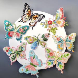 May include: A collection of embroidered butterfly patches in various colors, including blue, pink, orange, and green. Each butterfly features intricate floral details and is arranged on a white circular surface.