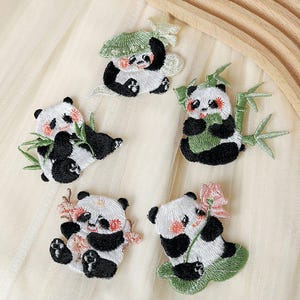 May include: Five embroidered panda bear patches with different designs. The patches are on a white fabric background. The pandas are black and white with green bamboo and pink flowers.