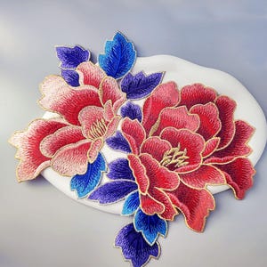 Puede incluir: Aplicación floral bordada con dos grandes flores de peonía en tonos rojos y rosas, con hojas azules y moradas. Las flores tienen detalles dorados y están sobre un fondo blanco de forma ovalada.
