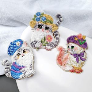 May include: Three embroidered patches of adorable cats wearing hats. One cat wears a yellow straw hat with blue flowers, another a blue beret, and the third a purple hat. Each cat has a unique design and color scheme.
