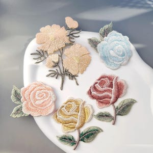 Puede incluir: Parches florales bordados en varios colores, incluyendo melocotón, azul claro, rosa, amarillo y burdeos. Los parches presentan costuras detalladas y hojas verdes, dispuestos sobre una superficie blanca. Ideal para la decoración textil.