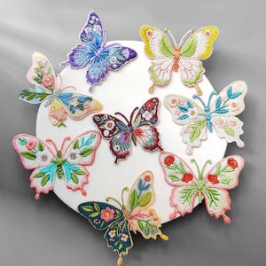 Puede incluir: Una colección de ocho parches de mariposas bordadas en varios colores, incluyendo azul, verde, rosa y amarillo. Cada mariposa presenta patrones florales detallados y costuras intrincadas, exhibidas sobre una superficie circular blanca.