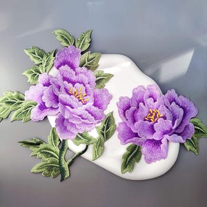 May include: Embroidered purple peony flower patches with green leaves and stems. The flowers have yellow centres and are set against a white, curved background. These patches are ideal for fabric embellishment.