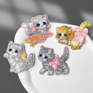 Puede incluir: Cuatro parches bordados termoadhesivos con gatitos de grandes ojos azules. Un gatito es gris con un lazo rosa, otro es marrón dorado con un lazo rosa, otro es gris con un lazo amarillo y otro es gris oscuro con un lazo rosa en la espalda.