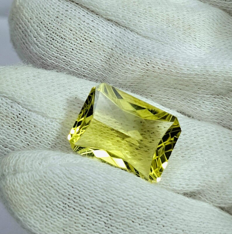 Pode incluir: Uma pedra preciosa facetada, de corte retangular, com um tom amarelo p&aacute;lido. A gema &eacute; segurada em um tecido branco, mostrando sua clareza e a precis&atilde;o de seu corte. As bordas da gema s&atilde;o bem definidas, refletindo a luz.