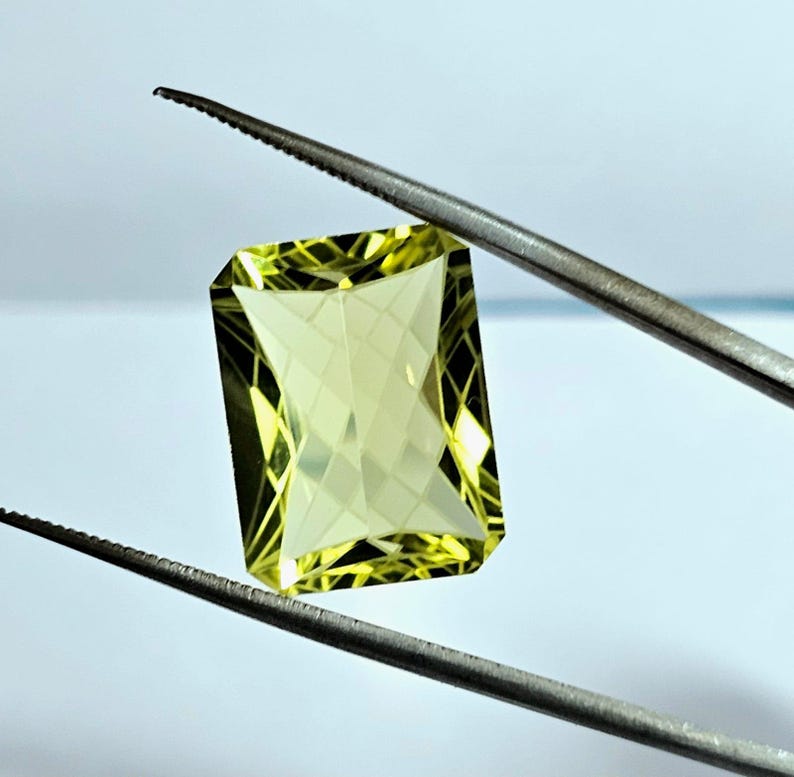 Pode incluir: Uma gema facetada, de corte retangular, em tom amarelo p&aacute;lido &eacute; segurada por uma pin&ccedil;a. As bordas da gema s&atilde;o bem definidas e seu interior reflete a luz, criando um efeito brilhante. A pin&ccedil;a &eacute; prateada.