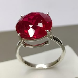 Può includere: Un anello d'argento con una grande gemma rosso scuro, taglio ovale. La gemma è tenuta da quattro griffe, e la fascia è in argento lucido. L'anello è esposto su una superficie bianca.
