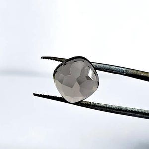 Puede incluir: Una gema de cuarzo ahumado facetada en forma de corazón, sujeta con unas pinzas. La gema es de color gris ahumado translúcido y tiene una superficie pulida. Las pinzas son de metal y tienen puntas dentadas. El fondo es blanco liso.