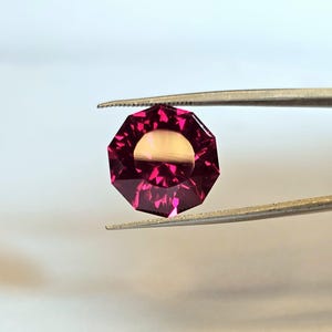 May include: A faceted, octagonal gemstone held by tweezers. The gem is a deep magenta color with a lighter, golden center. The facets catch the light, creating a sparkling effect. The gem is likely for jewelry making.