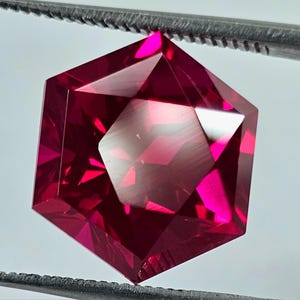 May include: A hexagonal-cut gemstone in a deep red colour, held by tweezers. The gem's facets create multiple reflections, highlighting its sharp angles. The stone's colour varies from a dark crimson to a bright, almost neon, pink.